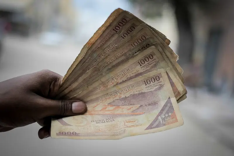 Une main tient en éventail une liasse de billets de 1 000 shillings somaliens usés, sur fond de rue floue.