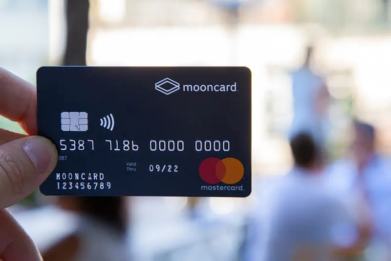 Une main tient devant l'objectif une carte bancaire noire Mooncard Mastercard, avec un arrière-plan flou où l'on devine des personnes attablées.